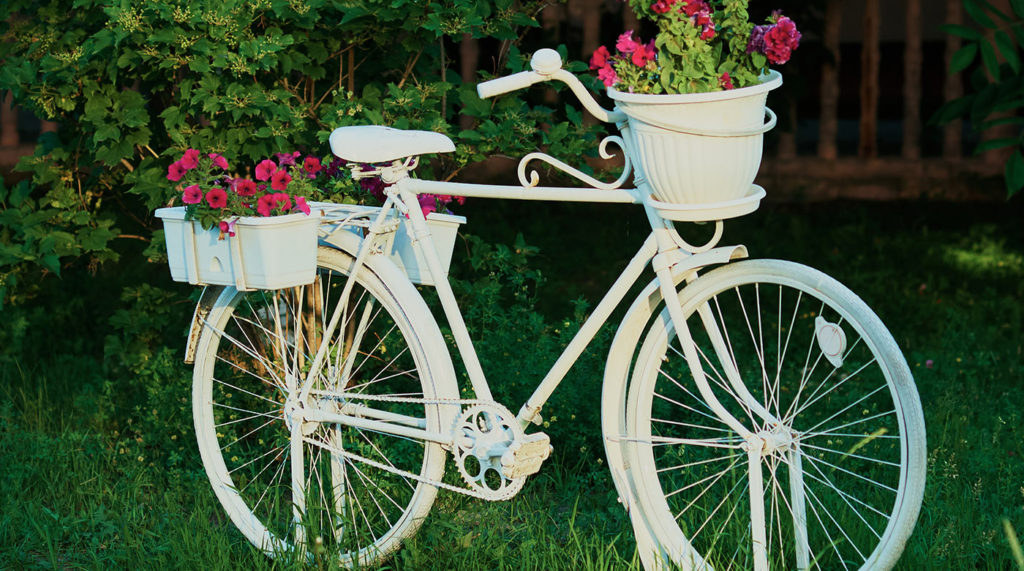 Weißes Deko-Fahrrad mit im Grünen mit pinken Blumen auf dem Gepäckträger und im Frontkorb. Für das Ehrenamt-Team zusammen ankommen. Foto; AdobeStock